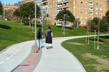 two girls walk uphill on a concrete track in the park towards the residential buildings