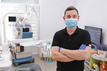 Fototapeta na wymiar A male dentist in uniform and medical face mask poses against a background of dental equipment in a dental office. Healthy teeth and medicine concept. dentist's reception