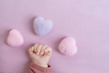 Tiny baby girl hand surrounded by three heart shape wool felt balls on light pink background.