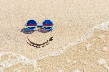 Fototapeta na wymiar A painted smile on the sand and sunglasses with the flag of the Cape Verde. The concept of a positive and successful holiday in the resort of the Cape Verde.