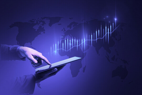 Close up of businessman hand pointing at tablet with absytract glowing business chart and map hologram on blue background. Finance, trade and analysis concept.