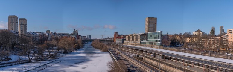Fototapeta premium Panorama, water front buildings, iicy canal Karlbergskanalen, traffic route Karlbergsleden, train tracks, dome and tower buildings, bridge St Eriksbron a sunny day in Stockholm