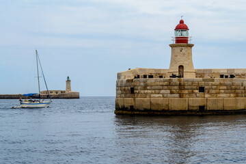 Fototapeta na wymiar A sailing boat passing between the Ricasoli Breakwater lighthouse & Valletta Breakwater lighthouse at the entrance to the Grand Harbour in Malta