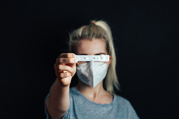 blond women with FFP-2 mask holding a test kit for viral disease COVID-19 2019-nCoV with a positive result in front of a black background.