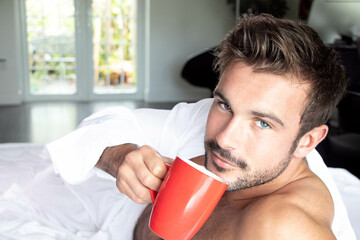 Sexy, naked muscular man with beard lying in bed covered with sheet drinking coffee