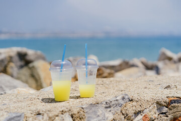 Colorful fresh drinks in plastic transparent glasses with blue straws on sandy rocks background with sea and sky on the horizon