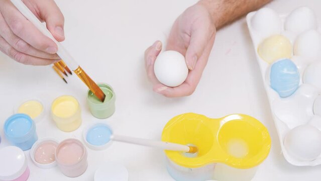 Close up view of man painting Easter eggs with special brush. Holiday concept. Guy paints an Easter egg green. Preparing for Easter. White table, top view.