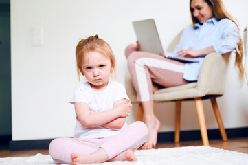 Fototapeta premium Upset little girl sitting lonesome while mother using laptop. Mother don't have time for daughter