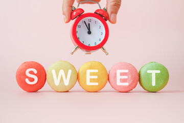 hand holding red analog alarm clock above sweet word  on multicolor macarons  with sweet pink...