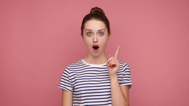 Overjoyed happy young adult woman pointing finger up looking at camera with toothy smile, having idea, plan, startup, wearing striped T-shirt. Indoor studio shot isolated on pink background.