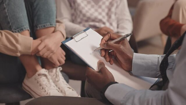 Slowmo closeup of unrecognizable African-American therapist taking notes on clipboard while having group therapy session with teenage kids talking about their problems