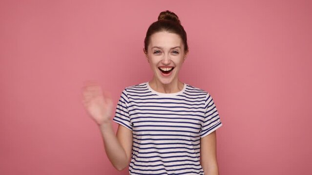 Hello. Adorable friendly beautiful woman turning to camera waving hand with engaging toothy smile, saying hi, wearing striped T-shirt. Indoor studio shot isolated on pink background.