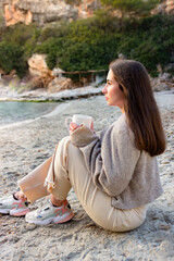 Young woman drinking from a mug, sitting in the sand facing the sea, enjoying the sunset views of the natural landscape with spring clothes. Concept of enjoying coffee or tea.