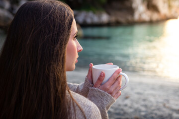 Woman drinking from a mug, sitting in the sand facing the sea, enjoying the sunset views of the natural landscape. Concept of enjoying coffee or tea.