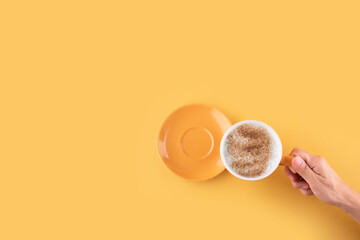 hand holding cup of cappuchino coffee on a yellow background. Space for text.