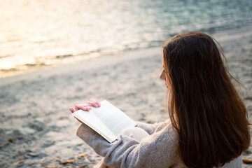 Relaxed unrecognizable woman sitting on the sand reading a book on a beach in spring clothes.