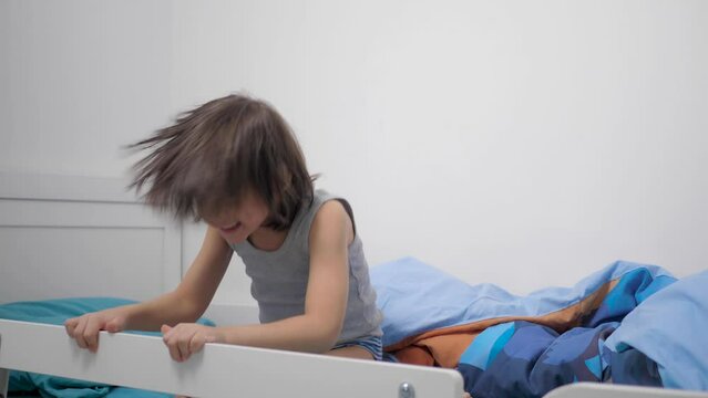 boy 5 years with long hair and a gray T-shirt is raging in his bed before going to bed and does not want to go to bed. hiding under a blanket, pillow and dancing