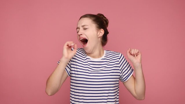 Portrait of sleepless woman yawning and raising hands up, feeling fatigued, wakes up early in the morning, wearing striped T-shirt. Indoor studio shot isolated on pink background.