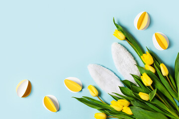 Yellow tulips, paper crafting eggs and white fluffy rabbit ears on blue background, easter and spring concept