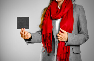 A girl in grey coat and red scarf holds a black clean empty square sticker. Isolated on grey background