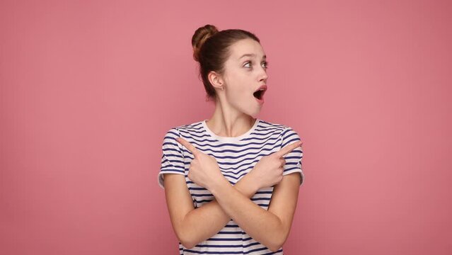 Side view of pleasantly surprised woman standing with crossed hands and pointing to copy space on both sides, wearing striped T-shirt. Indoor studio shot isolated on pink background.
