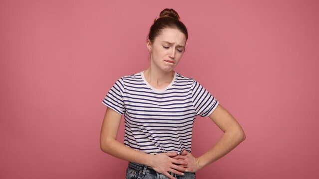 Upset woman holding her belly, suffering from terrible acute pain, symptoms of appendicitis inflammation, risk of miscarriage, wearing striped T-shirt. Indoor studio shot isolated on pink background.