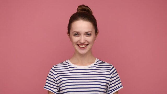 Happy optimistic attractive woman with hair bun nodding yes, expressing agreement, looking at camera with positive expression, wearing striped T-shirt. Indoor studio shot isolated on pink background.