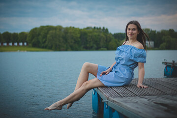 Fototapeta na wymiar Cute brunette model with long straight dark hair in a summer blue dress with a beautiful realistic smile poses by a lake in the Czech Republic. European woman. Candid portrait with realistic smile