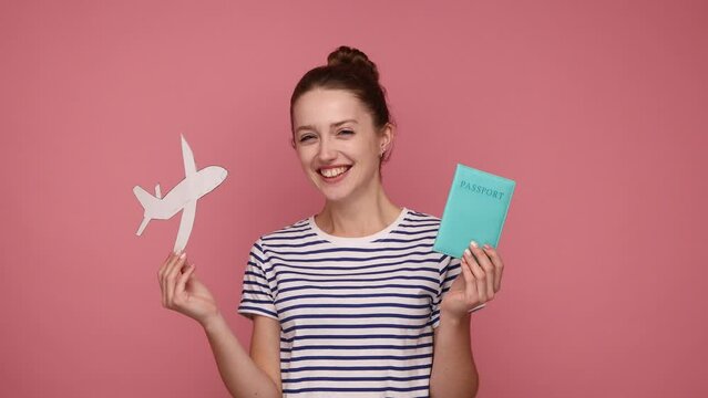 Extremely happy excited woman with bun hairstyle holding paper plane and passport, being glad she has a vacation, wearing striped T-shirt. Indoor studio shot isolated on pink background.