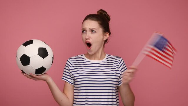 Extremely happy woman holding in hands flag of USA and soccer ball, supporting favourite team on championship, rejoicing, wearing striped T-shirt. Indoor studio shot isolated on pink background.