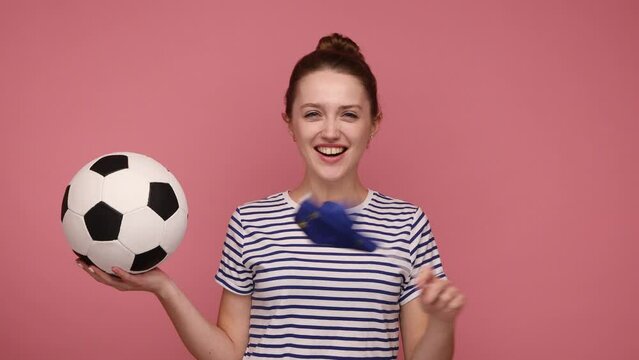 Active happy woman holding in hands flag of Europe and soccer ball, supporting favourite team on championship, rejoicing, wearing striped T-shirt. Indoor studio shot isolated on pink background.