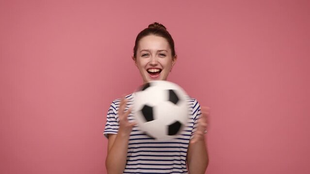 Happy attractive woman standing with soccer ball in hands and playing, being ready to go on football match and cheering, wearing striped T-shirt. Indoor studio shot isolated on pink background.