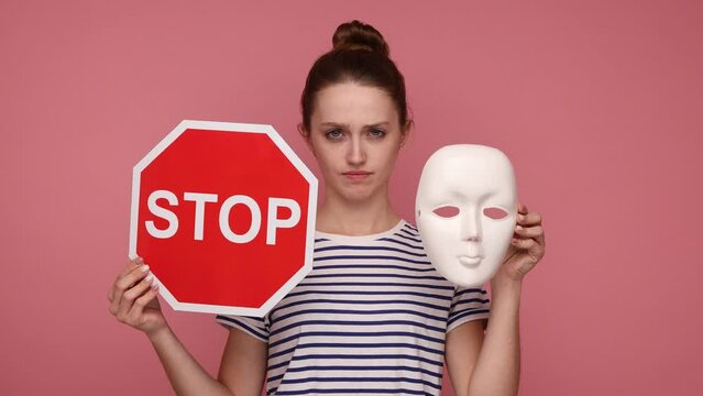 Woman holding white mask and stop traffic sign to camera, nodding her head no, don't change your personality, wearing striped T-shirt. Indoor studio shot isolated on pink background.
