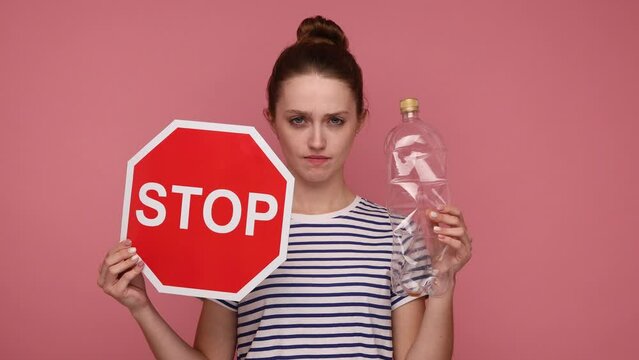 Strict serious woman with bun hairstyle holding red traffic style and empty plastic bottle, sorting your rubbish, reusing, wearing striped T-shirt. Indoor studio shot isolated on pink background.