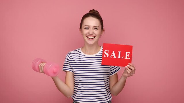 Successful smiling woman showing holding sale card and dumbbell, black Friday discounts on gym membership, wearing striped T-shirt. Indoor studio shot isolated on pink background.