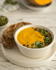 Close up pumpkin cream soup in a white bowl decorated with cesam seeds and cilantro nearby slice of bread