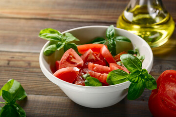 Fresh red tomatoes are sliced in a white ceramic plate with basil herb and olive oil on a wooden table.