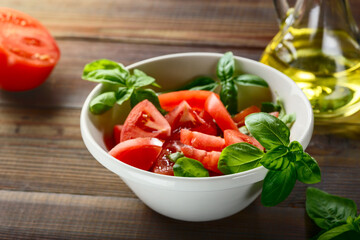 Fresh red tomatoes are sliced in a white ceramic plate with basil herb and olive oil on a wooden table.