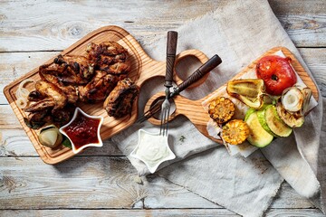 Hot barbecue Chicken wings on a cutting board and assorted grilled vegetables. Appetizing grilled chicken meat with vegetables and herbs, sauce. Top view. Copy space. Rustic wooden table background.