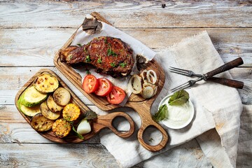 Grilled steak on a cutting board and assorted grilled vegetables. Appetizing grilled meat with vegetables and herbs, sauce. Top view