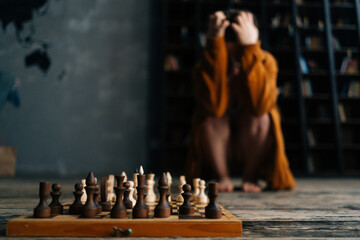 Close-up selective focus to chess board on wooden floor, worried unrecognizable woman sitting in...