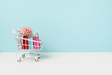 Trolley cart and colorful paper shopping bags with pink flower rose on blue background. Creative idea for shopping online, summer sale, supermarket, discount promotion. Copy space for text