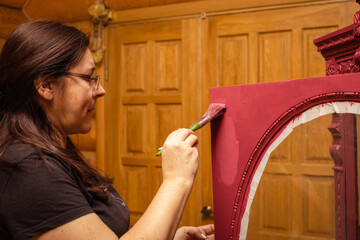Woman aging door of wood cupboard with nice carved ornaments by painting it in red color....