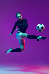 Obraz na płótnie Canvas Dynamic portrait of female soccer player practicing with football ball isolated on purple studio background in neon light. Sport, action, motion, fitness