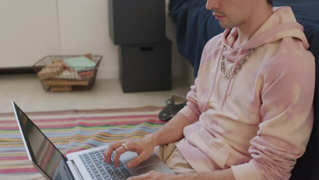 Tilting up of open-minded Caucasian male student wearing hoodie, sitting on floor in dorm room at daytime, using portable computer, typing