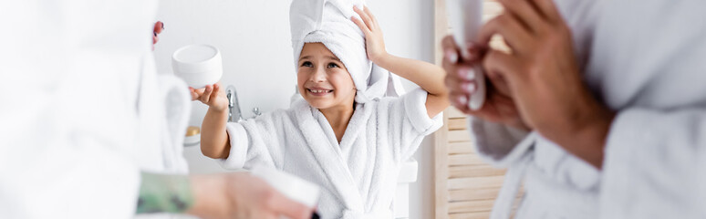 Smiling girl in bathrobe holding cream near blurred moms in bathroom, banner