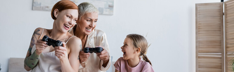 KYIV, UKRAINE - DECEMBER 8, 2021: Positive kid looking at mothers with joysticks at home, banner
