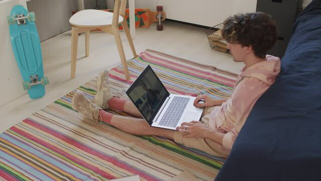 Side-view of curly-haired Caucasian male college student wearing hoodie, sitting on floor in dorm room at daytime, using portable computer, having remote lesson with teacher, talking