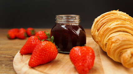 Strawberry jam in glass jar with fresh berries, croissant on wooden background