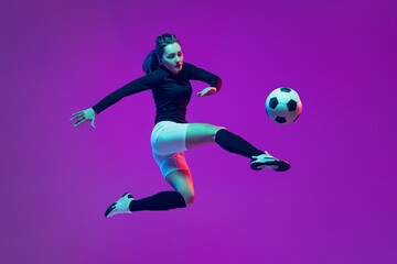 Obraz na płótnie Canvas One sportive girl, female soccer player kick in jump football ball isolated on purple studio background in neon light. Sport, action, motion, fitness